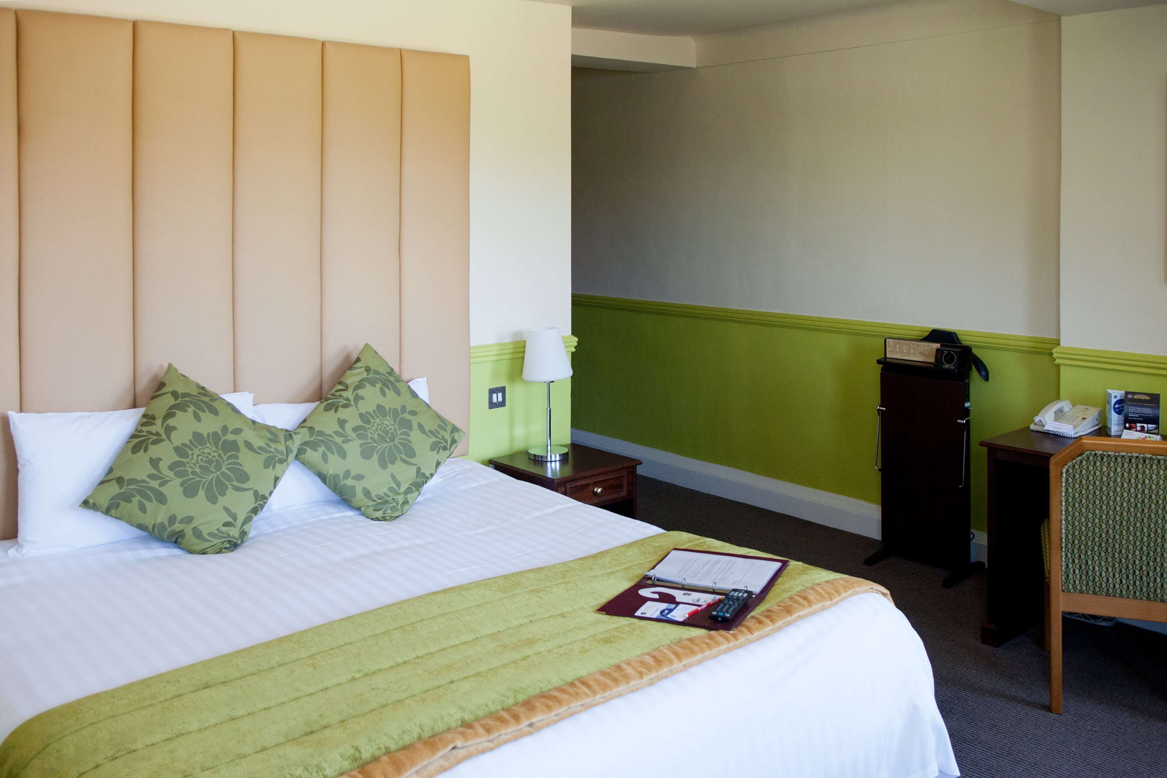 A hotel room with a bed, green and white bedding, a green throw blanket, a bedside table with a lamp, a telephone, and a small desk with a chair.