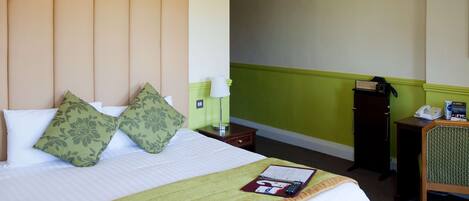 A hotel room with a bed, green and white bedding, a green throw blanket, a bedside table with a lamp, a telephone, and a small desk with a chair.