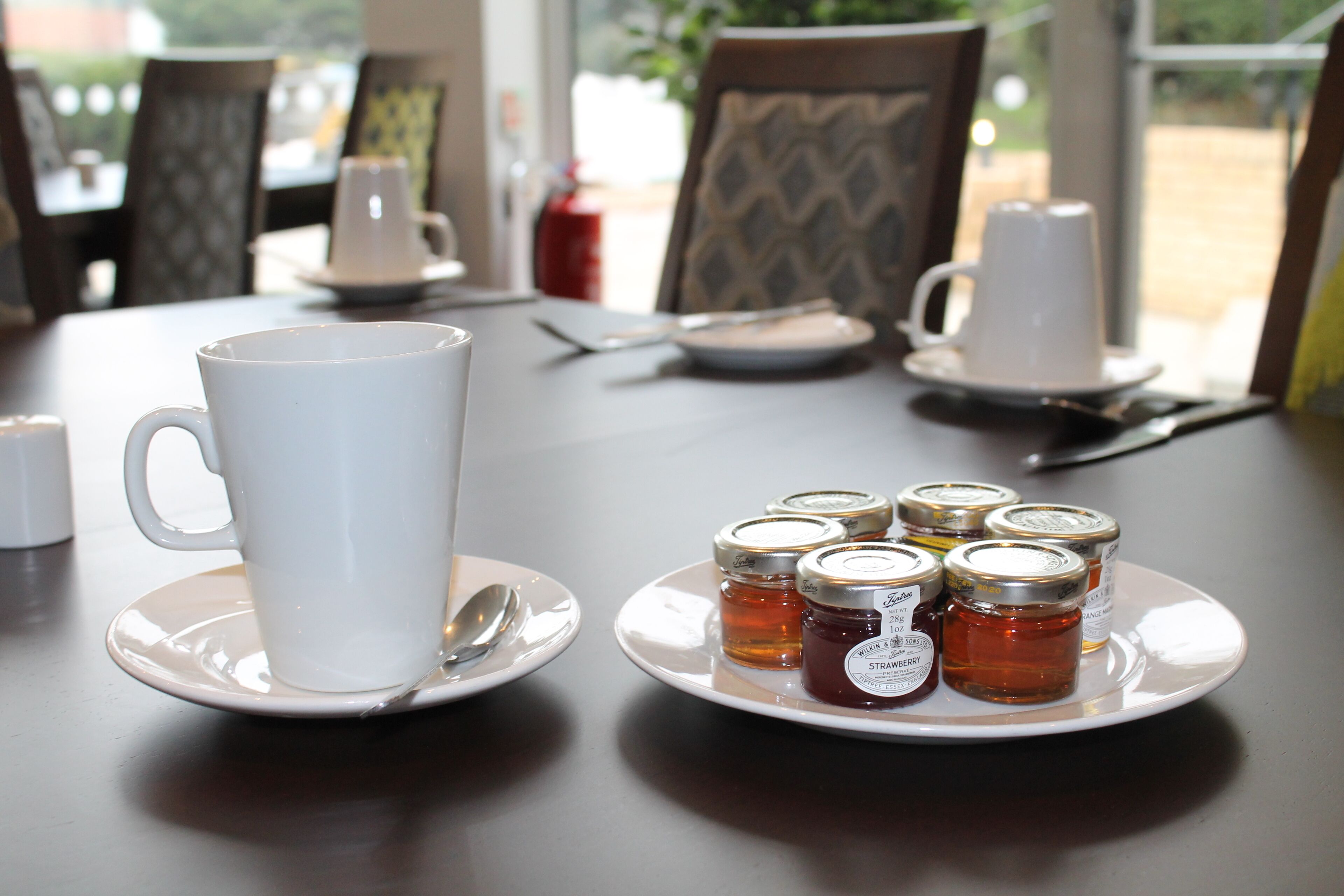 A table with a white cup, a plate with jam jars, and a spoon.