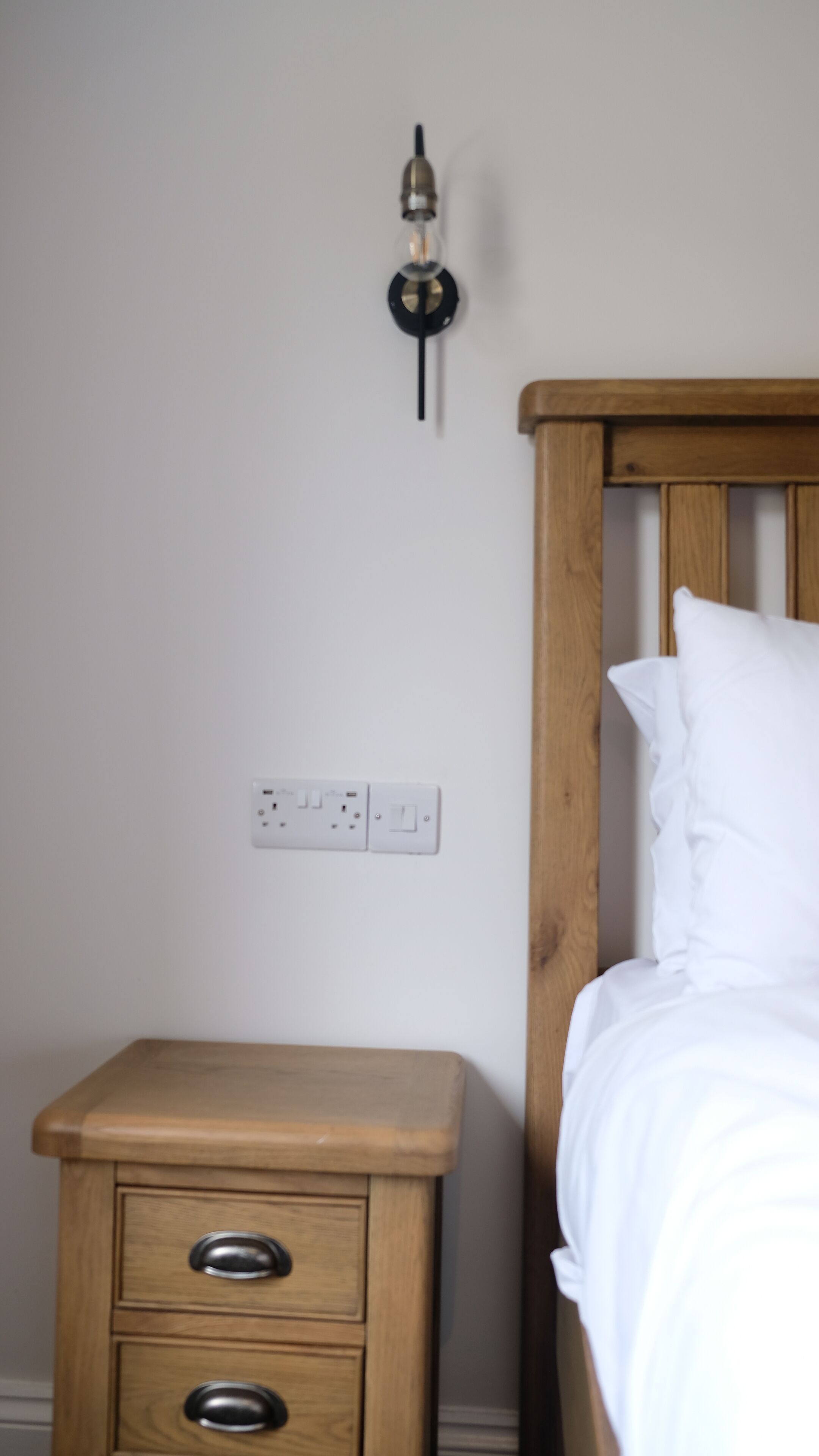 A wooden bedside table with drawers, a wall socket, and a light switch.