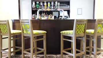 A bar area with a counter, multiple liquor bottles, glasses, and a computer monitor. There are four wooden chairs with yellow cushions.