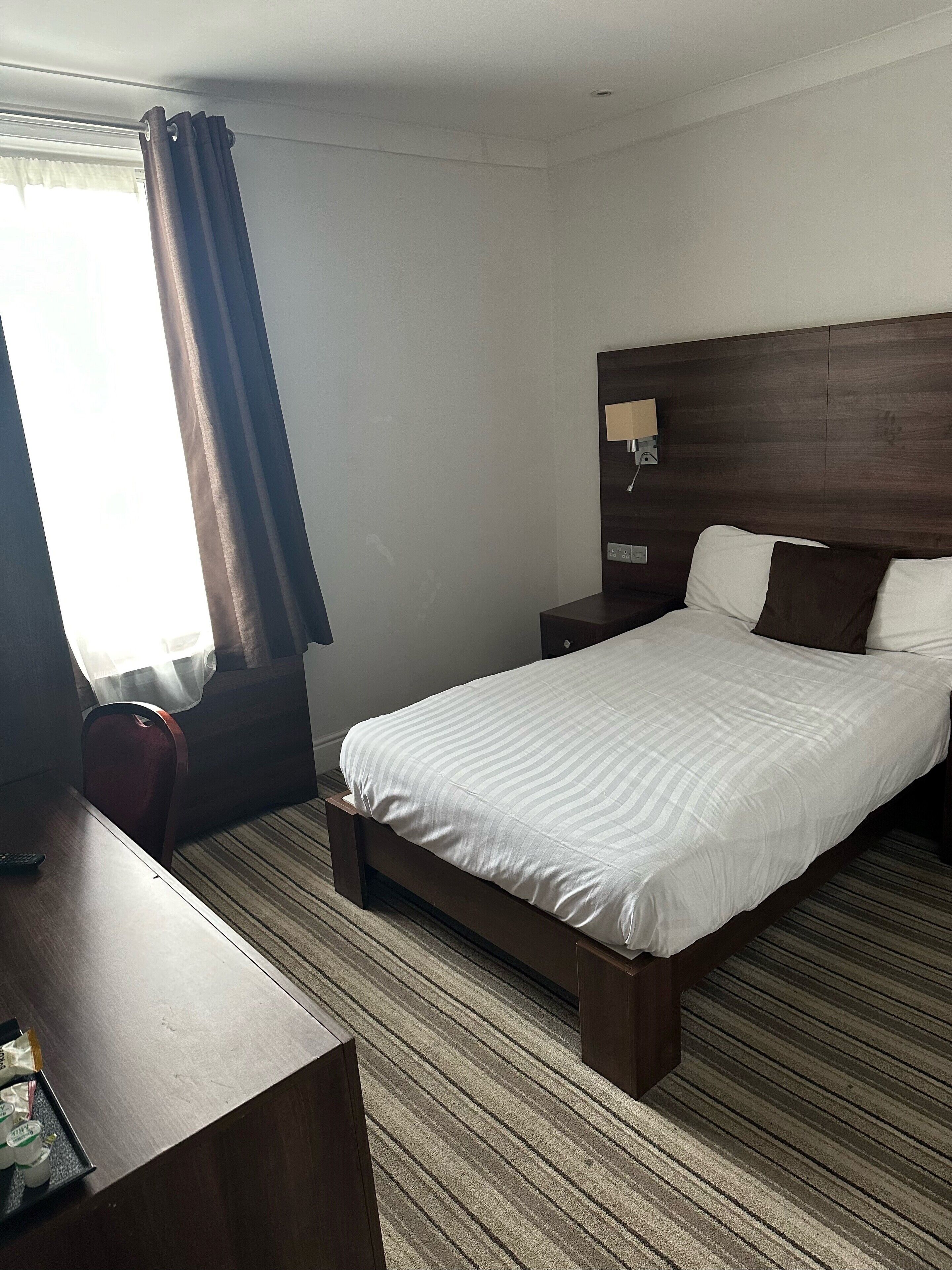 A hotel room with a bed, bedside table, lamp, and a view of the outdoors.