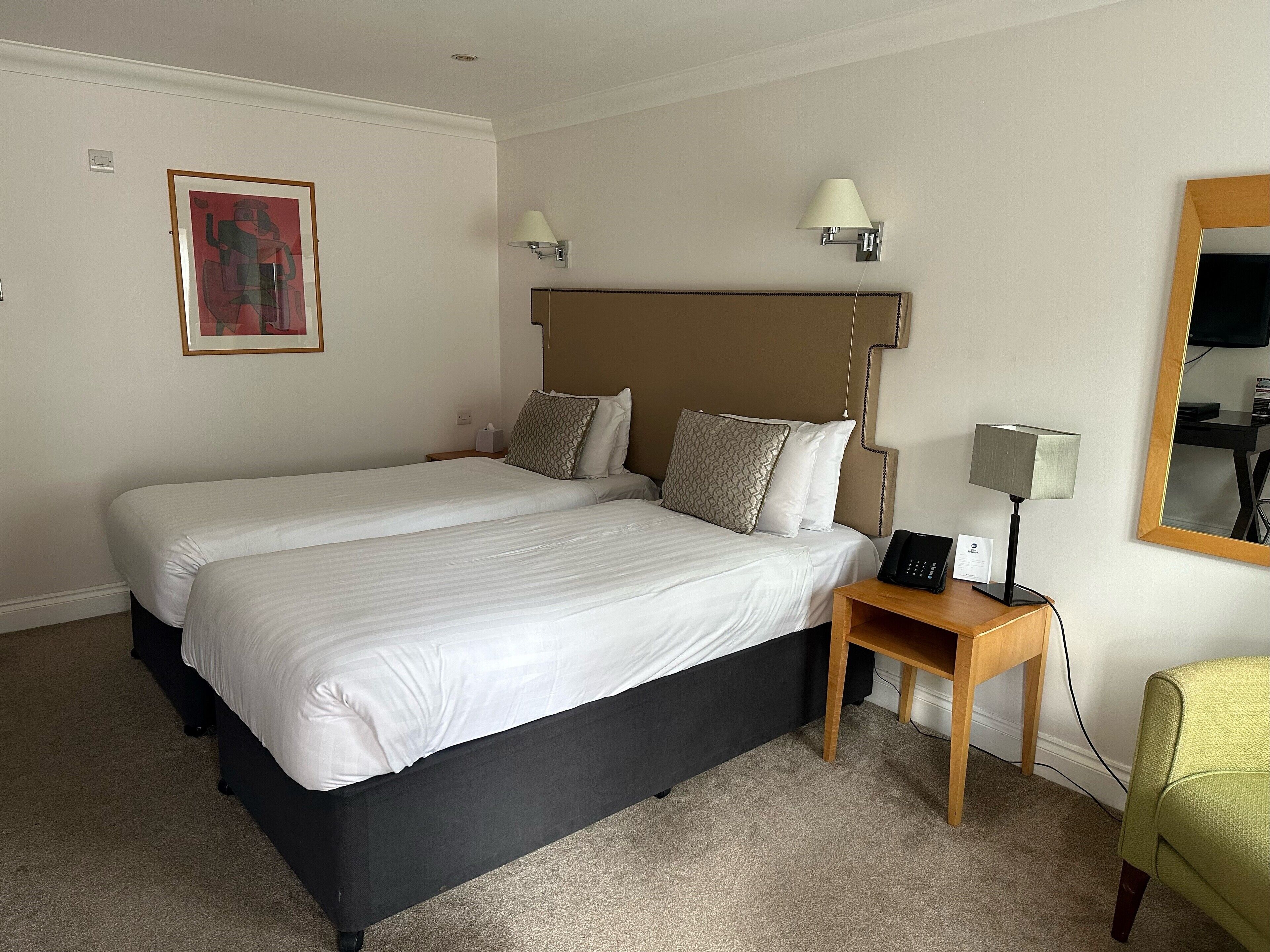 Quarto de hotel com duas camas, uma mesinha de cabeceira com abajur, telefone ao lado da cama e um quadro na parede. Espelho na parede oposta.