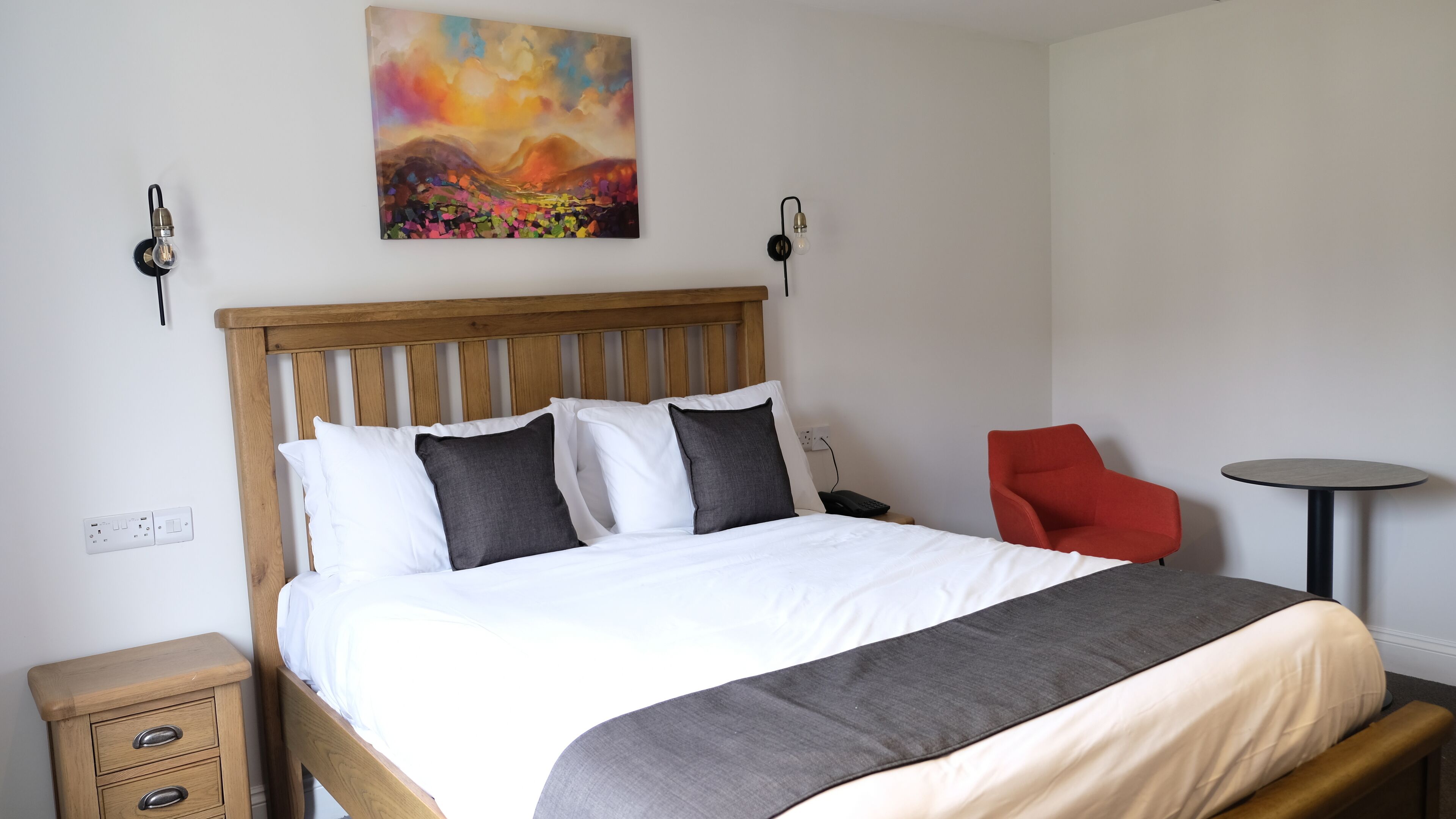 A neatly made bed with a wooden headboard, a bedside table with drawers, a red chair, and a small round table.