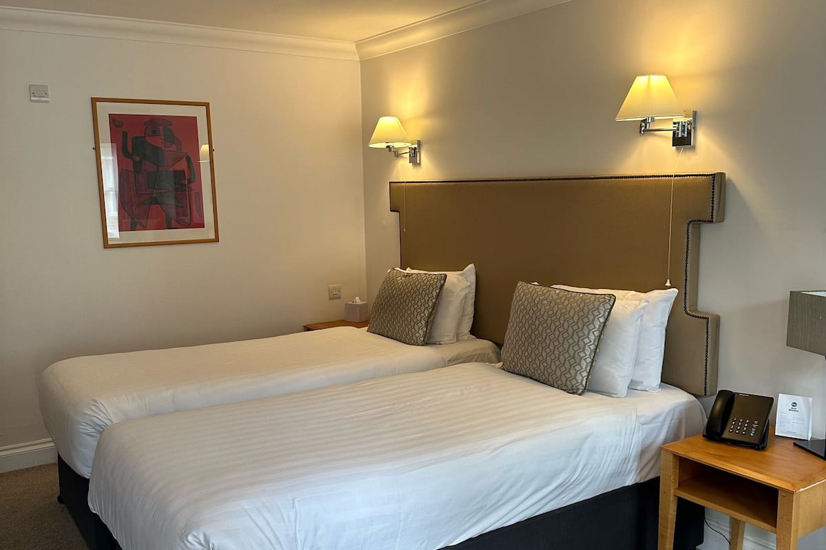 A hotel room with two beds, a headboard with integrated lighting, a bedside table with a phone and a lamp, and a framed artwork on the wall.
