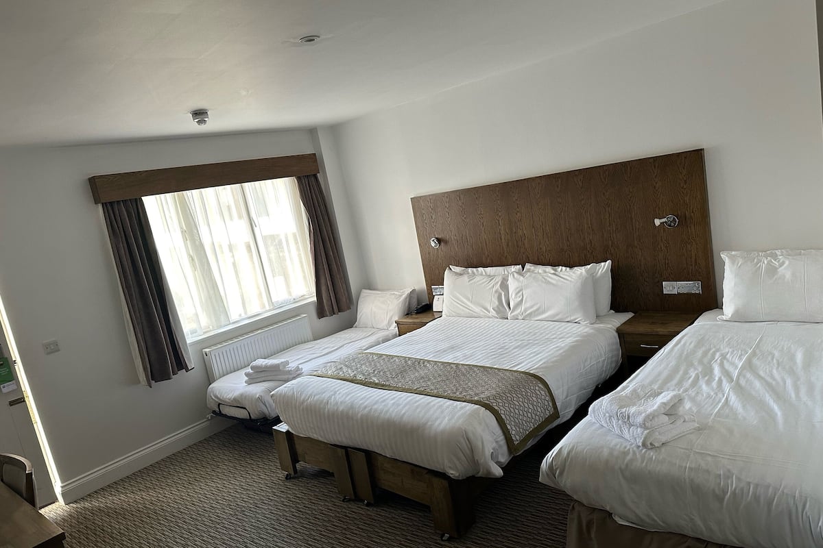 A hotel room with two beds, a wooden headboard, a window with curtains, a bedside table, and a radiator.