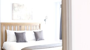 Ein ordentlich bezogenes Bett mit hölzernem Gestell und weißer Bettwäsche, eine Wandleuchte und ein Teilansicht eines Fensters mit Vorhängen.