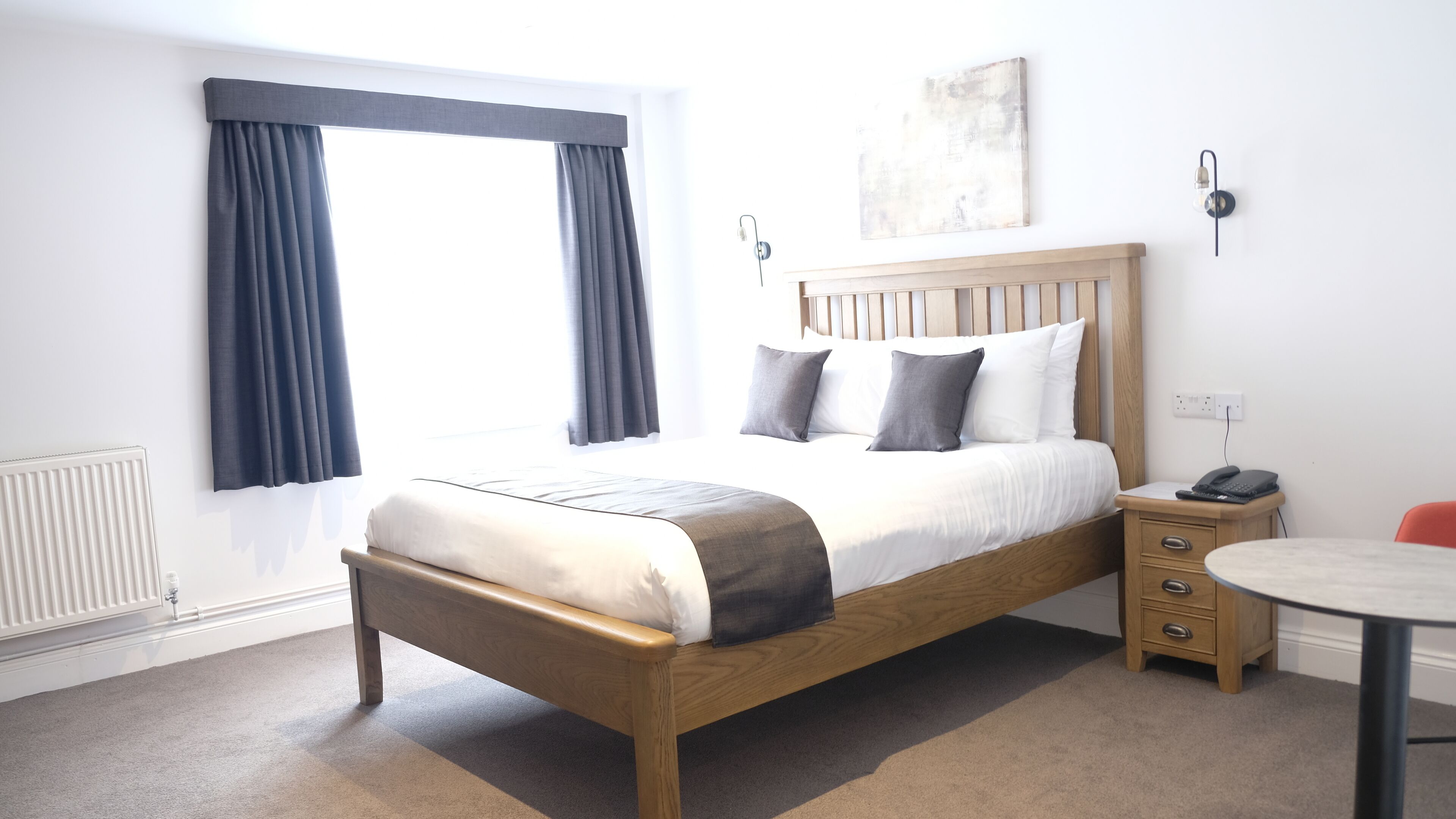 Un dormitorio ordenado con una cama de madera, una mesita de noche, una pequeña mesa redonda y un radiador.