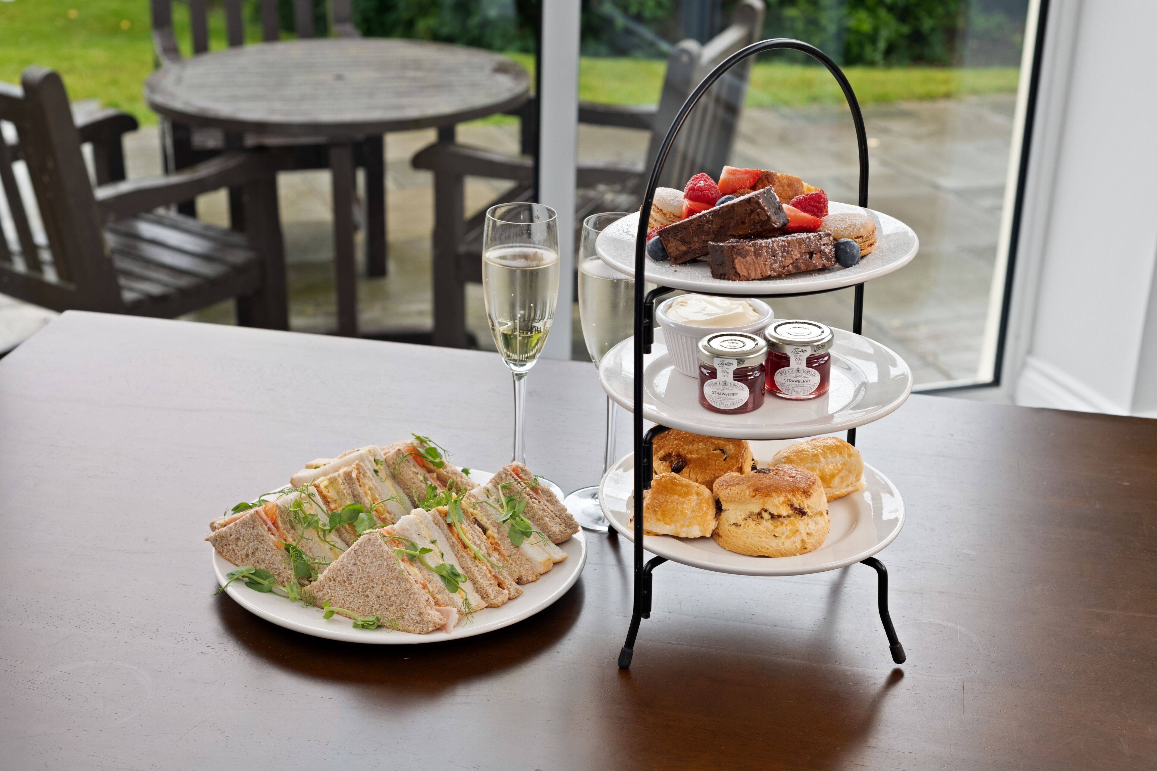A table with a three-tiered dessert stand, a plate of sandwiches, and a glass of white wine.