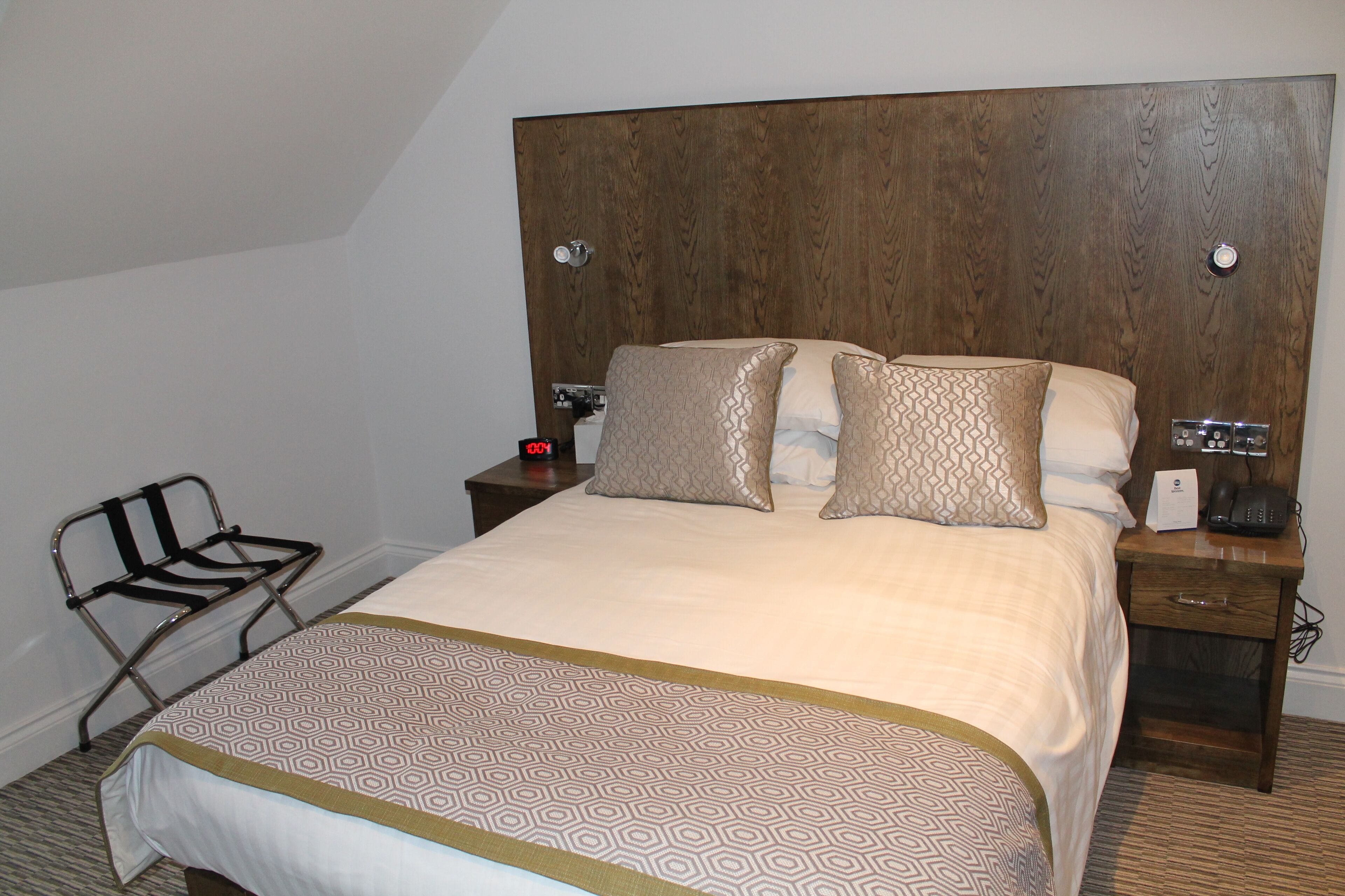 A neatly made bed with patterned bedding, a bedside table with a phone and a lamp, and a chair with a metal frame.