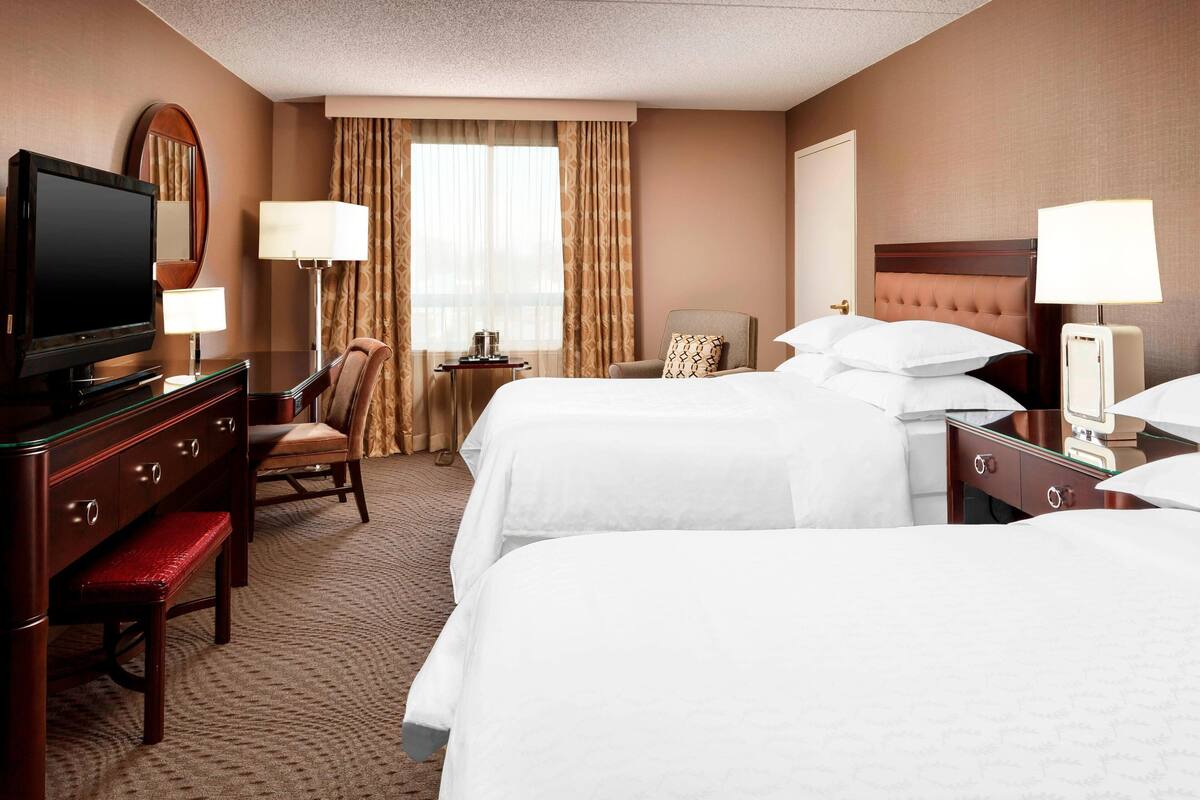 A hotel room with two beds, a desk with a television, a chair, a bedside table with a lamp, and a window with curtains.