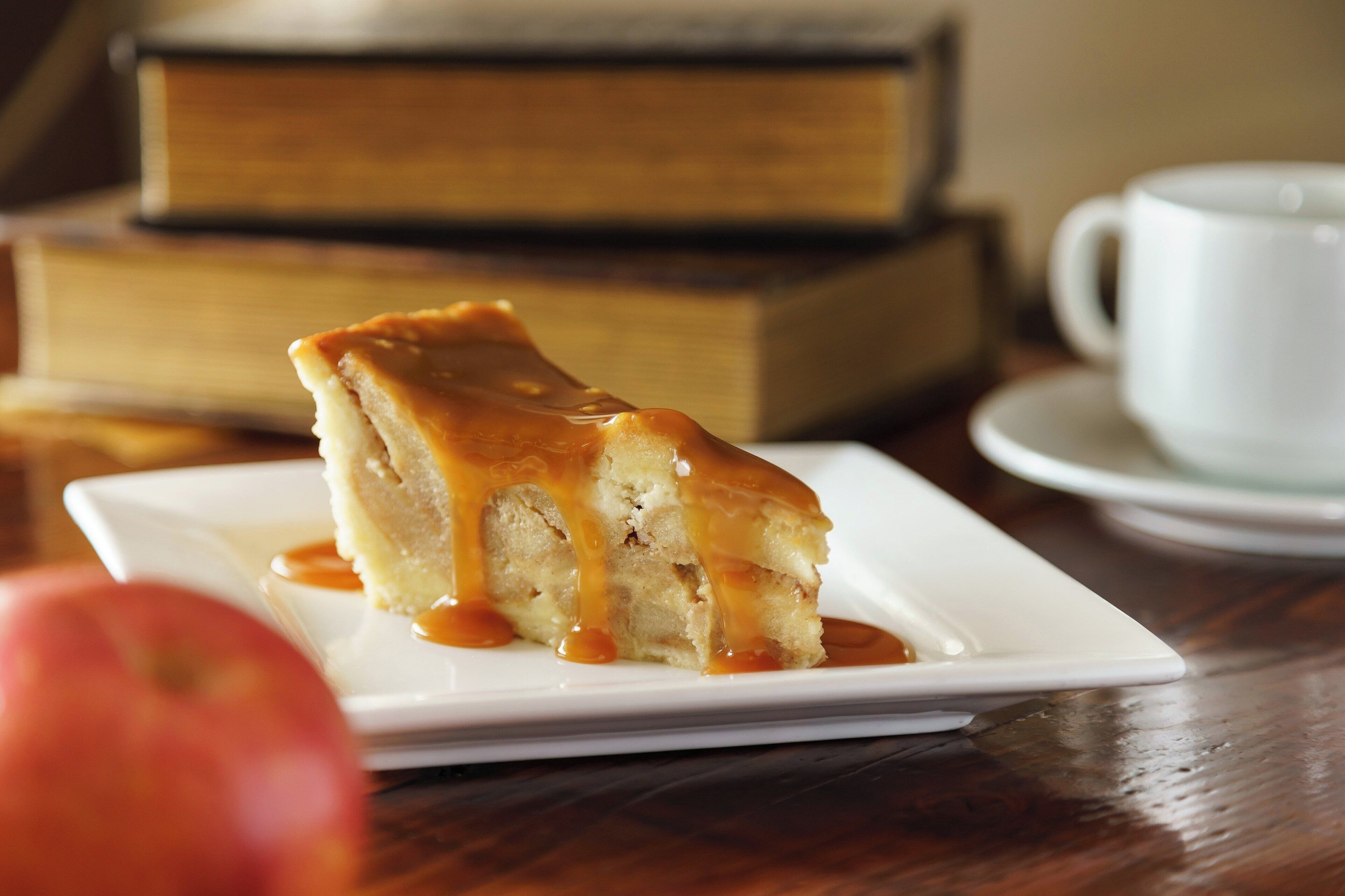Una porción de pastel con salsa de caramelo sobre un plato blanco, una taza blanca y una pila de libros al fondo.