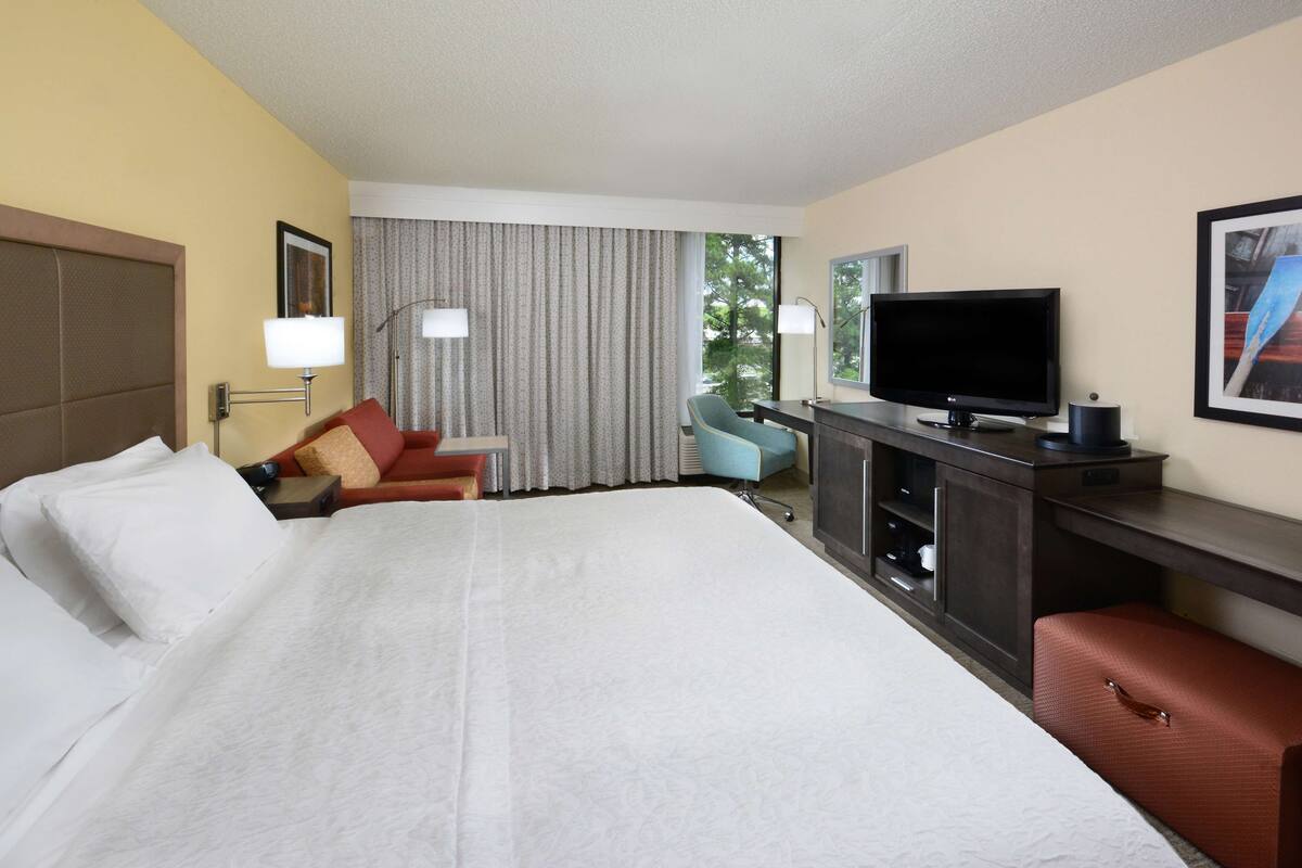 A hotel room with a large bed, a desk with a flat-screen TV, a chair, and a window with curtains.