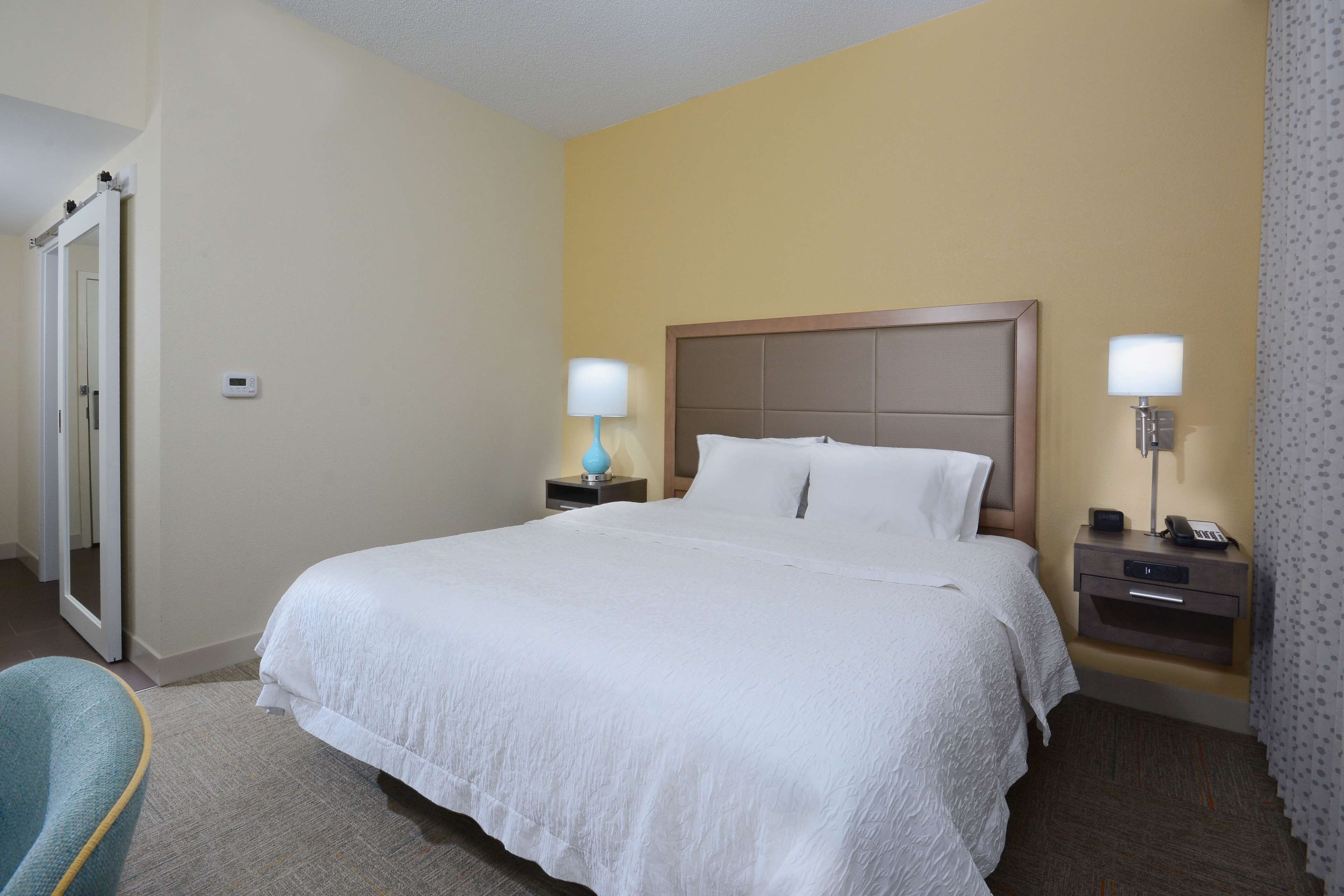 Quarto de hotel com uma cama grande, duas mesas de cabeceira com luminárias, um espelho e uma porta.