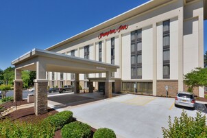 Un Hampton Inn con entrada cubierta, estacionamiento y cielo azul despejado.