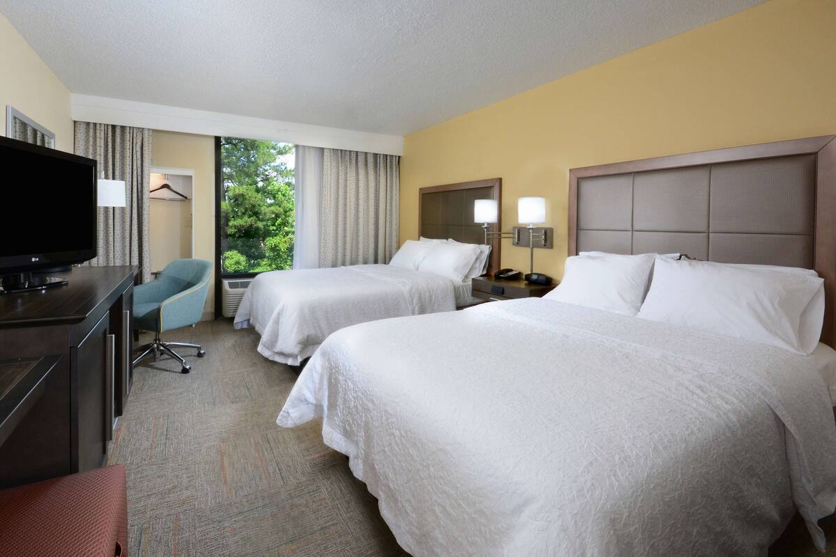 A hotel room with two beds, a desk with a television, a chair, and a window with curtains.