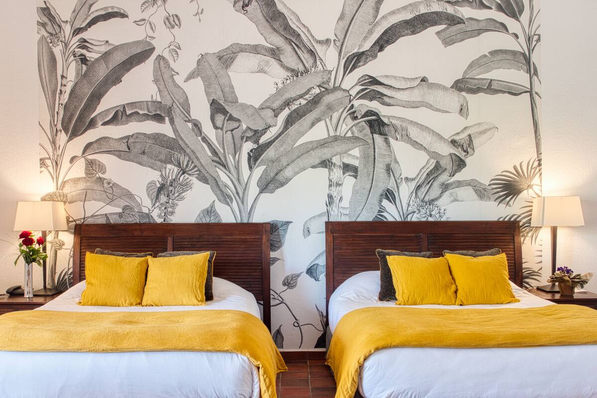 Ein Hotelzimmer mit zwei Betten, jeweils mit einem hölzernen Kopfteil, weißer Bettwäsche und gelben Kissen. An der Wand hinter den Betten befindet sich ein großes botanisches Wandbild.