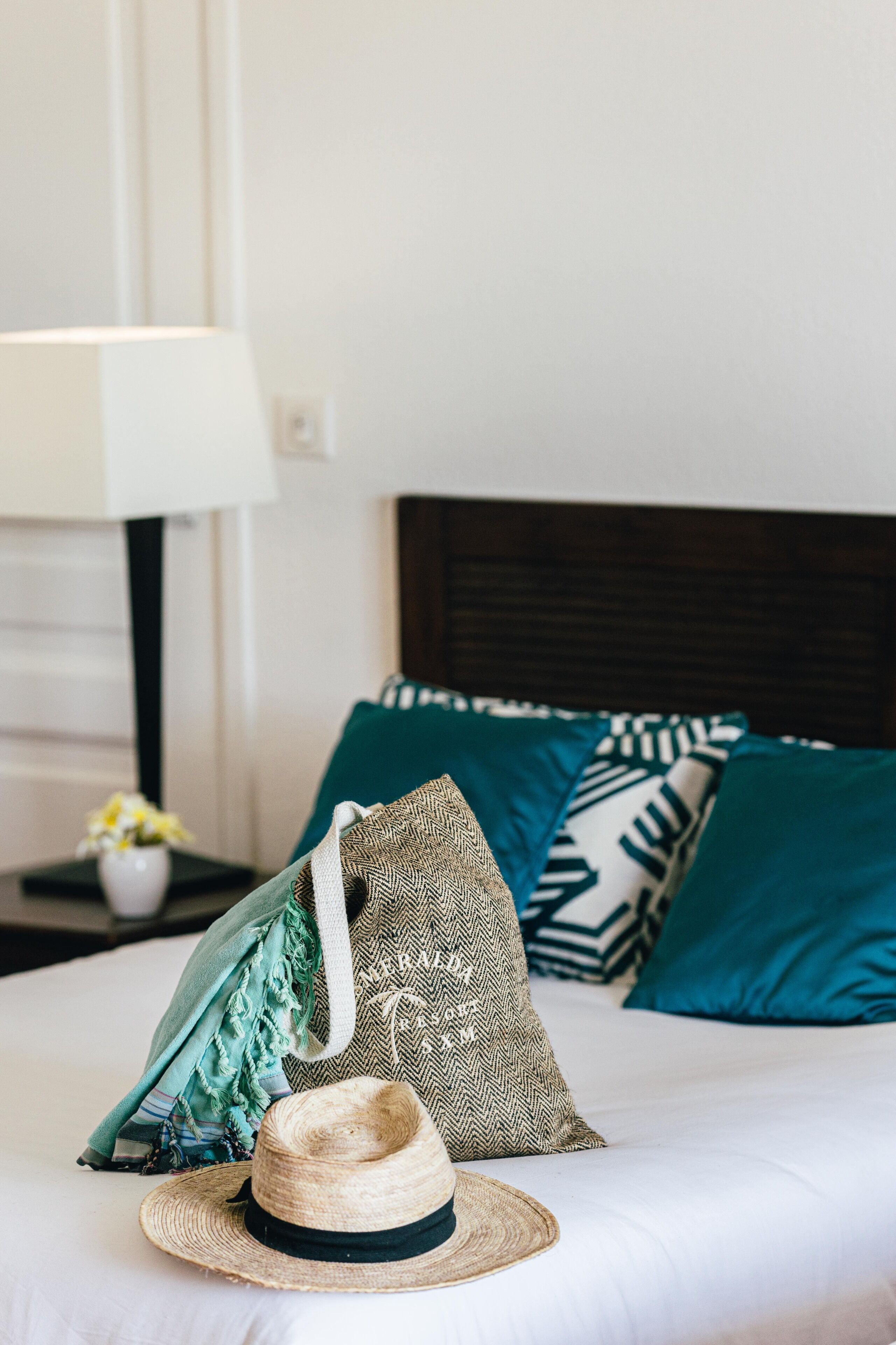 Una cama bien hecha con una colcha blanca, una almohada estampada en verde y blanco, y un sombrero de paja sobre la cama.