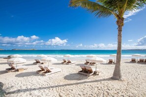 A beach with white lounge chairs and umbrellas, a palm tree, and a clear blue sky. - Esmeralda Resort (Orient Bay)