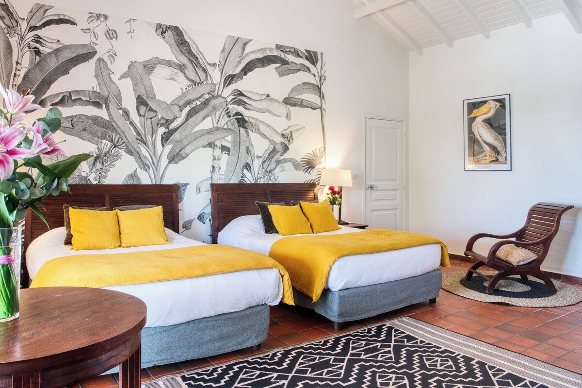 A hotel room with two beds, a large botanical wall mural, a wooden chair, and a patterned rug.