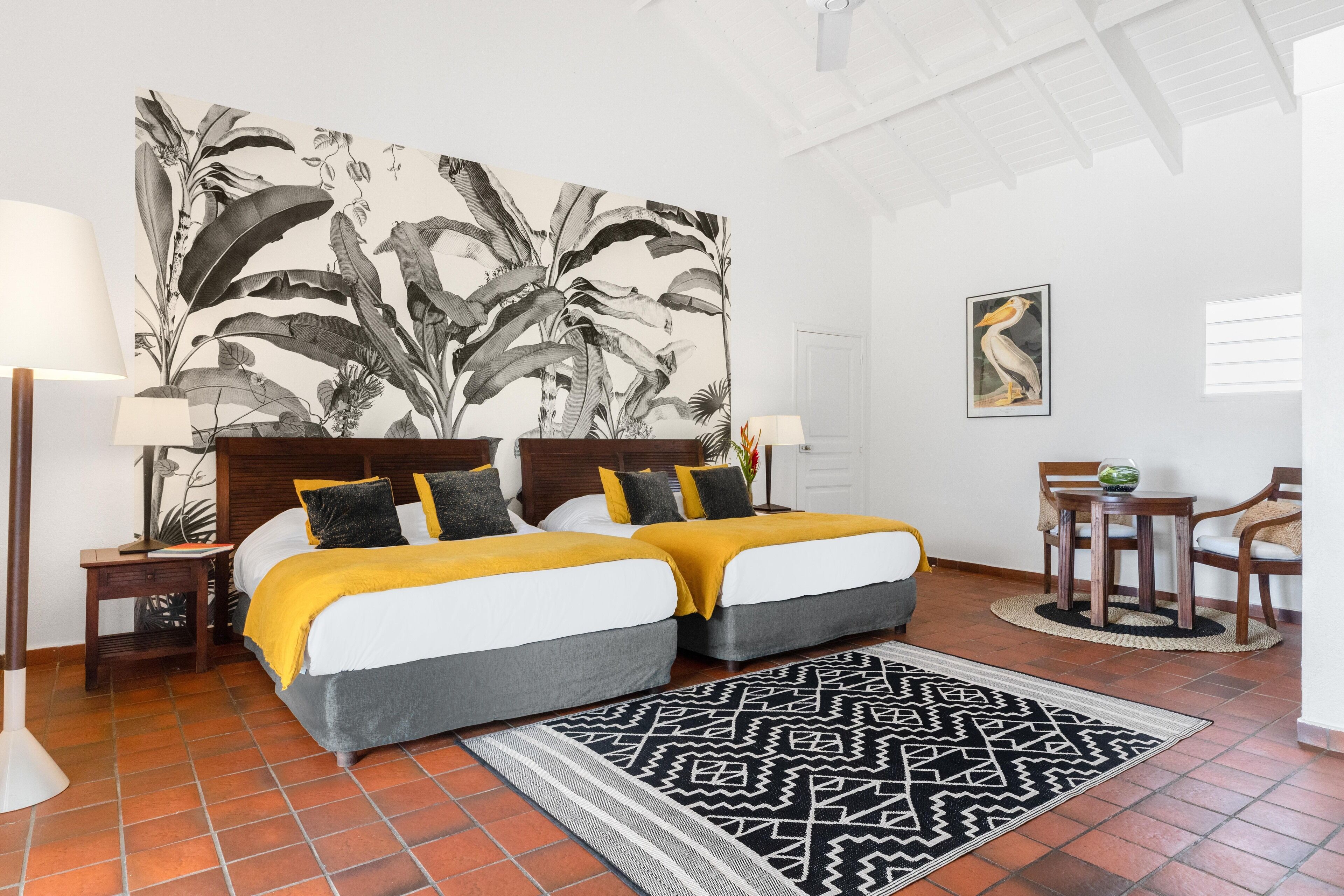 Una cama doble con colcha amarilla y gris, un tapete con estampado blanco y negro, y un cuadro de arte botánico de gran tamaño.