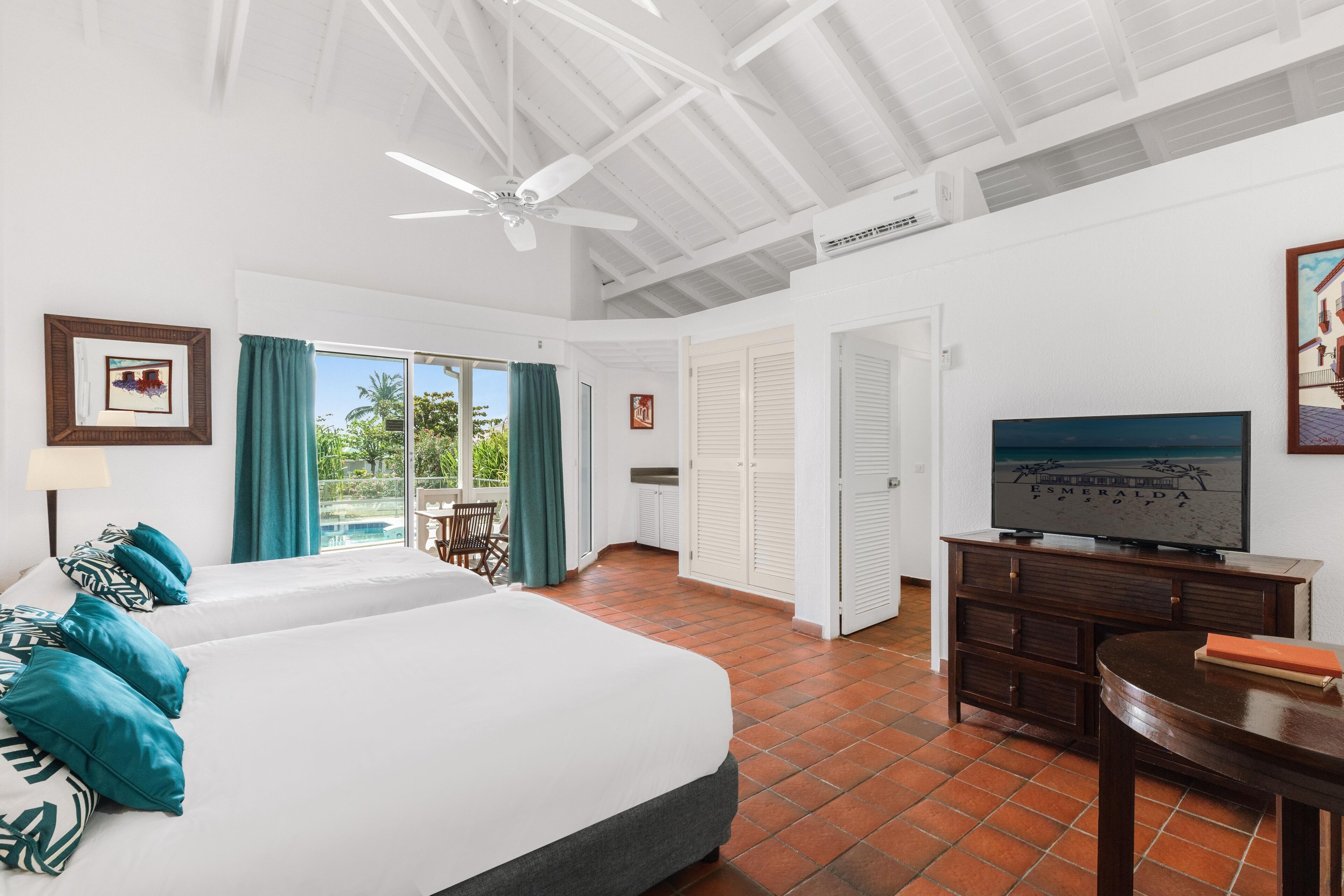A hotel room with a large bed, a TV, a desk, and a balcony with a view of greenery.