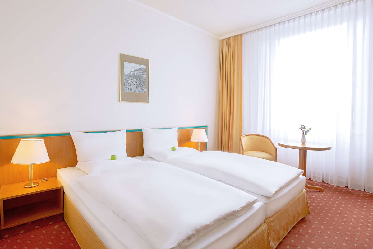 Habitación de hotel con dos camas, una mesita de noche, una silla, una pequeña mesa y una ventana con cortinas.