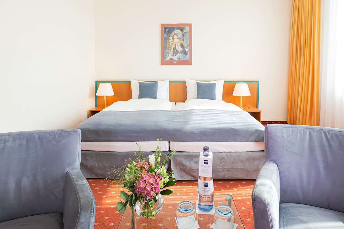Habitación de hotel con una cama grande, dos mesitas de noche con lámparas, un arreglo floral y una botella de agua sobre la mesita.
