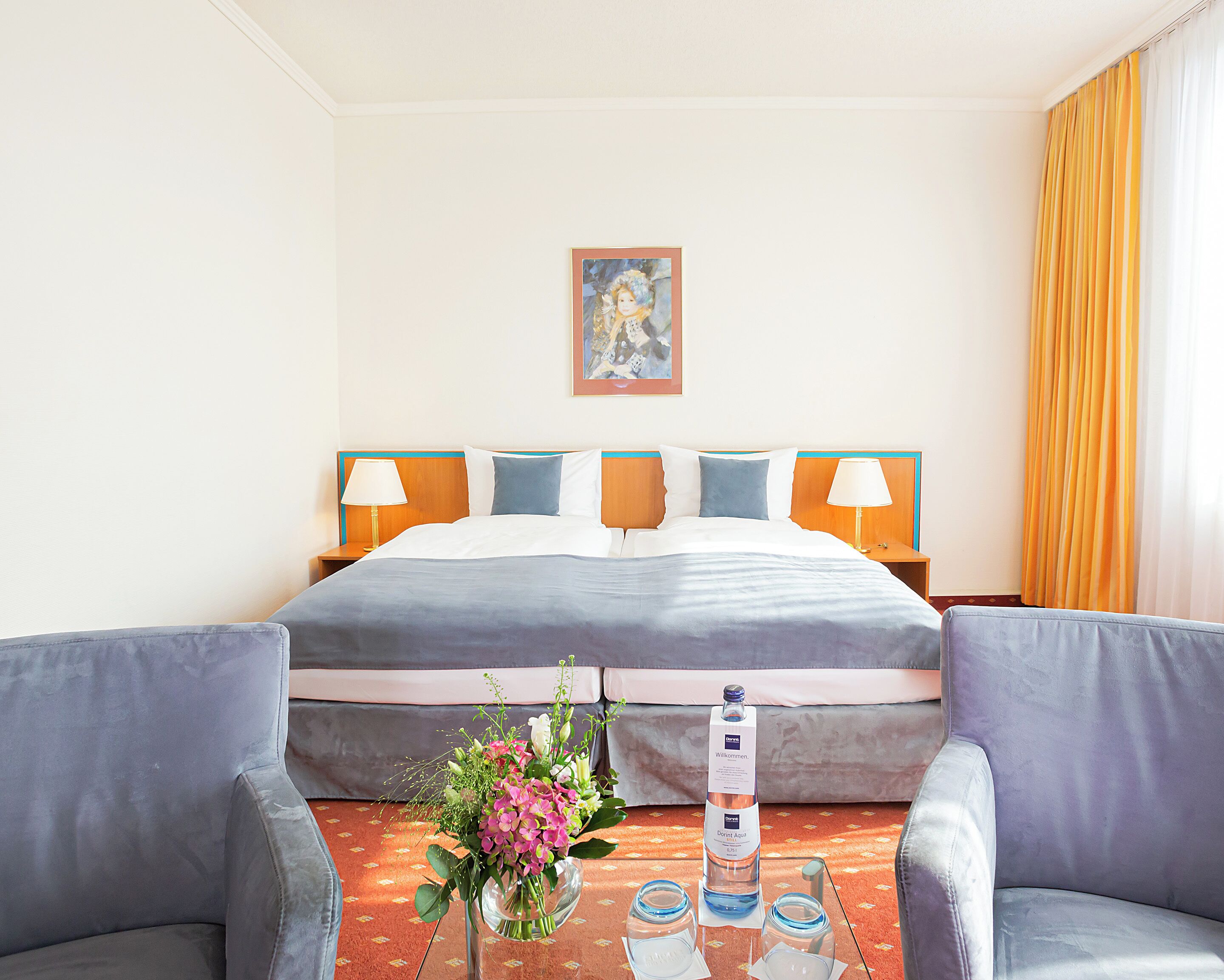 Habitación de hotel con cama, dos lámparas de noche, un cuadro en la pared, un arreglo floral y una botella de agua sobre la mesita de noche.