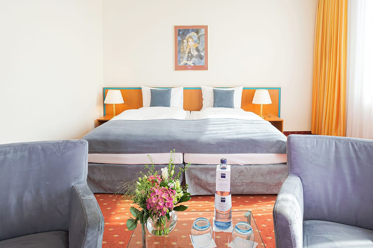 Habitación de hotel con cama, dos lámparas de noche, un cuadro en la pared, un arreglo floral y una botella de agua sobre la mesita de noche.