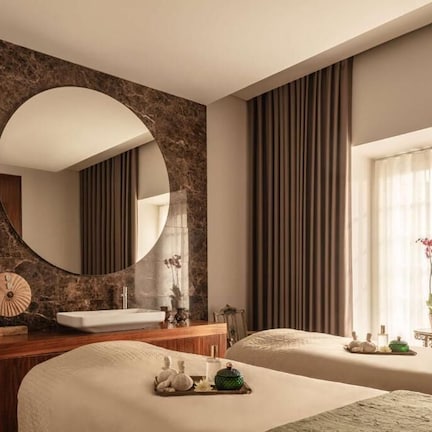 A hotel room with a marble wall, a round mirror, a bed with a tray of toiletries, a small table with a vase of flowers, and a window with curtains.