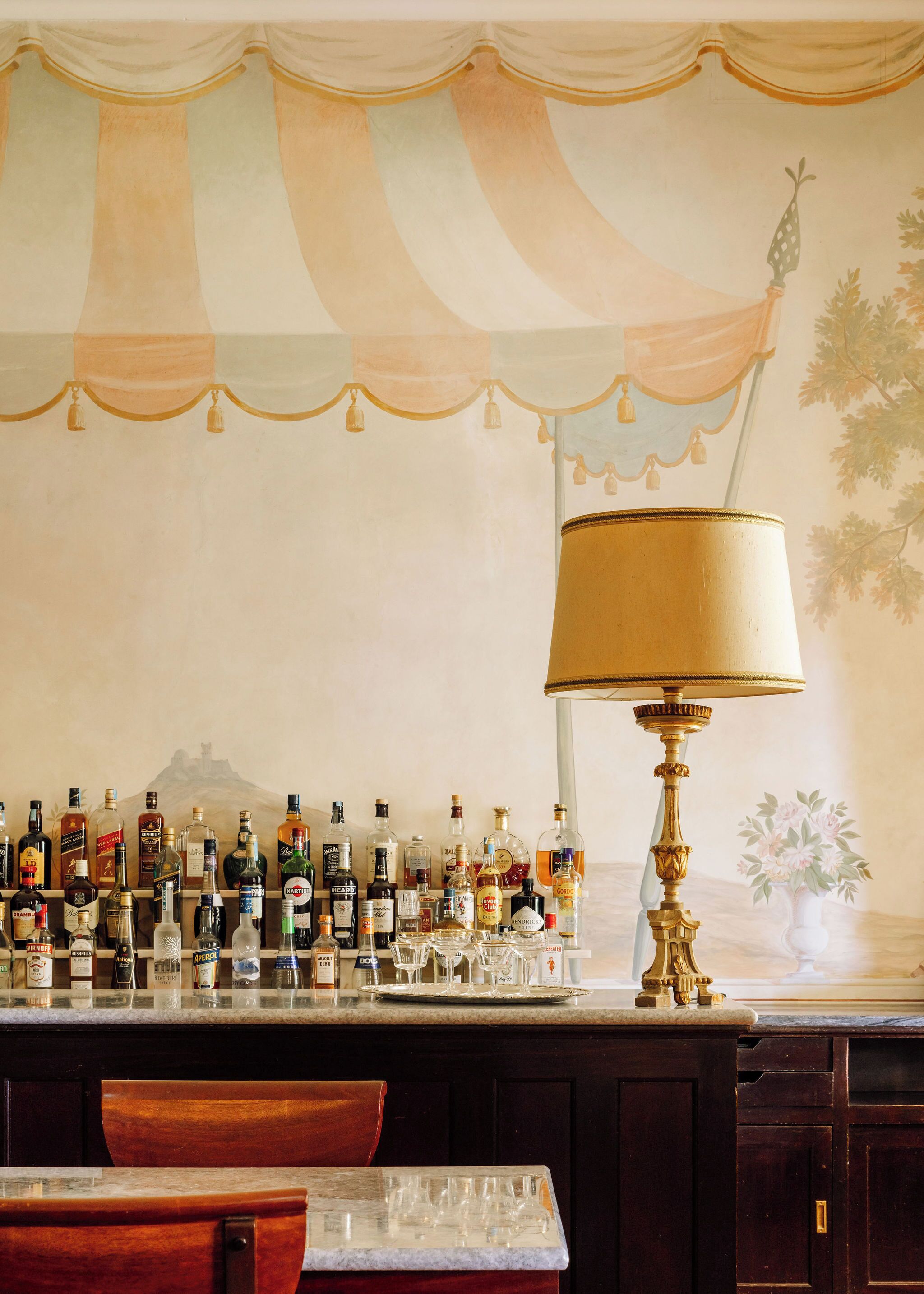 A bar area with a variety of liquor bottles and glasses, a decorative lamp, and a mural on the wall.