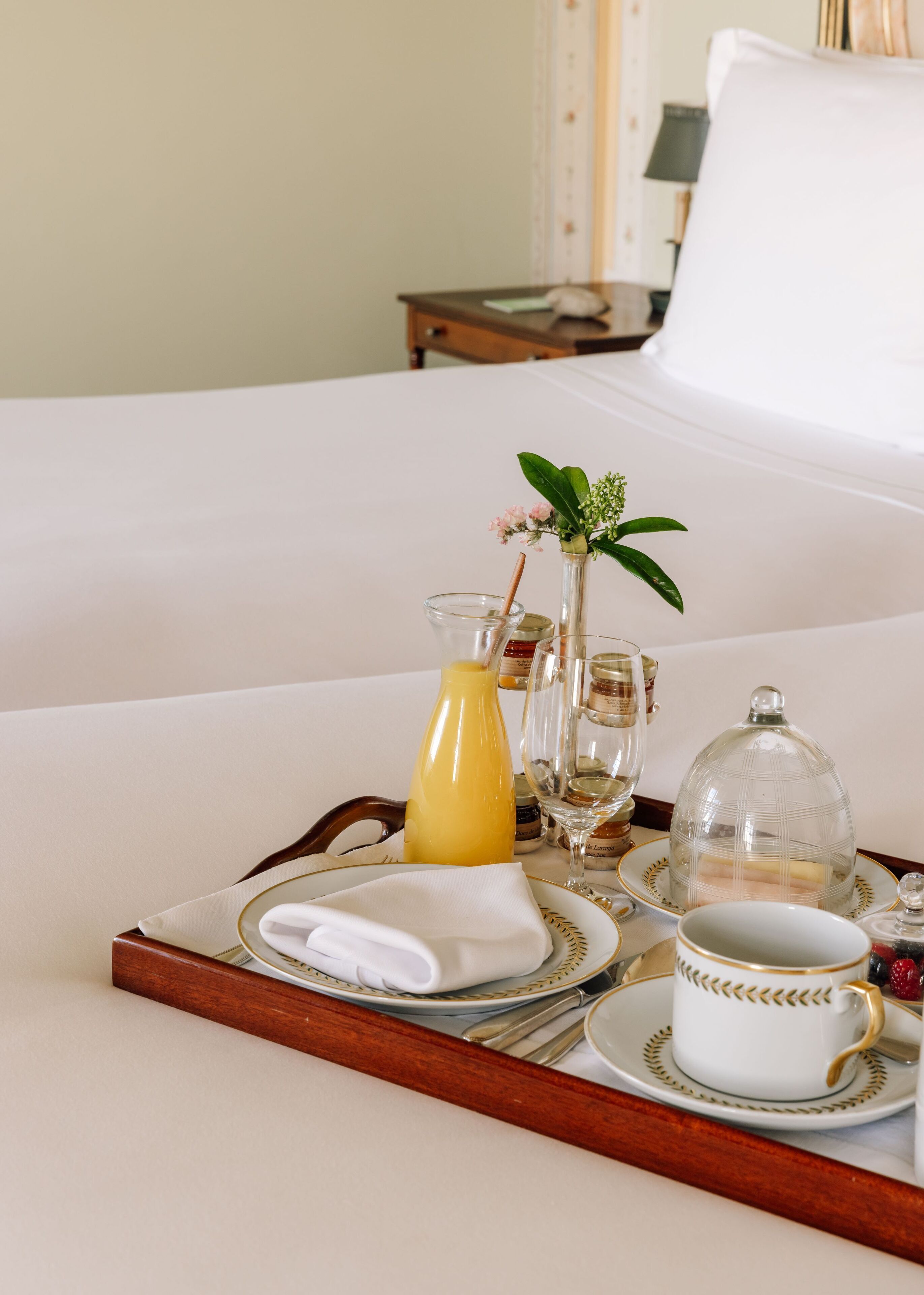 A breakfast tray with a glass of orange juice, a vase with flowers, a glass of water, a glass of milk, a cup of coffee, and a plate with a pastry on a bed.