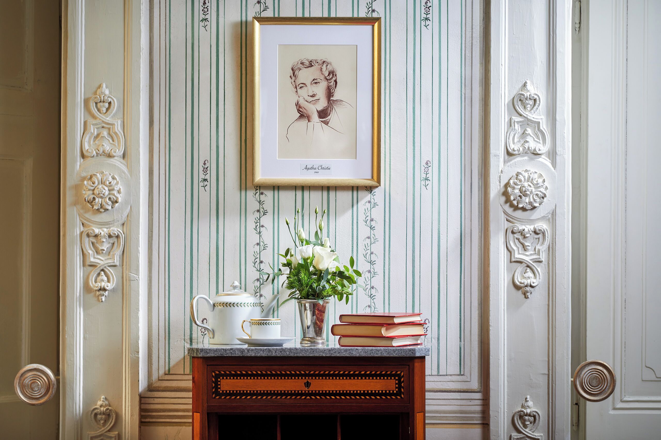 A framed portrait, a vase with flowers, a teapot, and a stack of books on a mantelpiece.