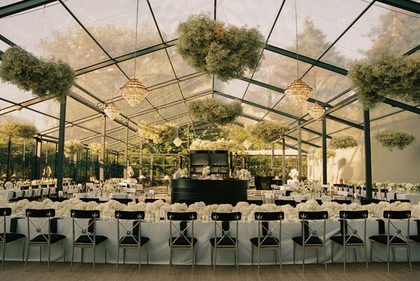 Un espacio tipo invernadero con mesas y sillas, candelabros y plantas colgantes.