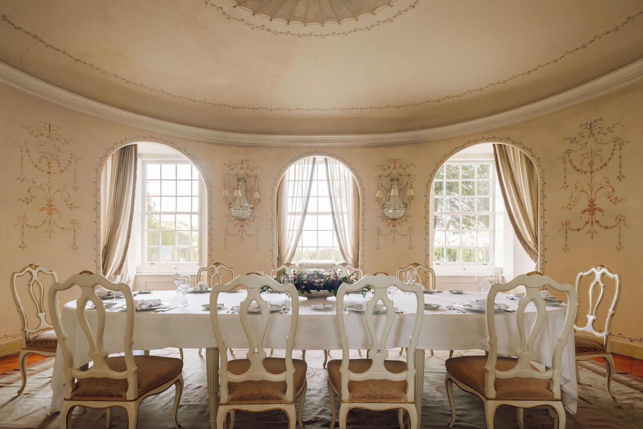 A formal dining room with a long table, ornate chairs, and three large windows with curtains.