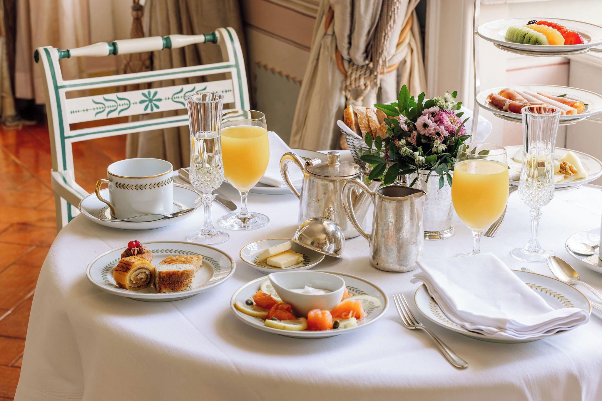 Una mesa de desayuno dispuesta con platos de comida, vasos de jugo de naranja, una jarra y un jarrón con flores.