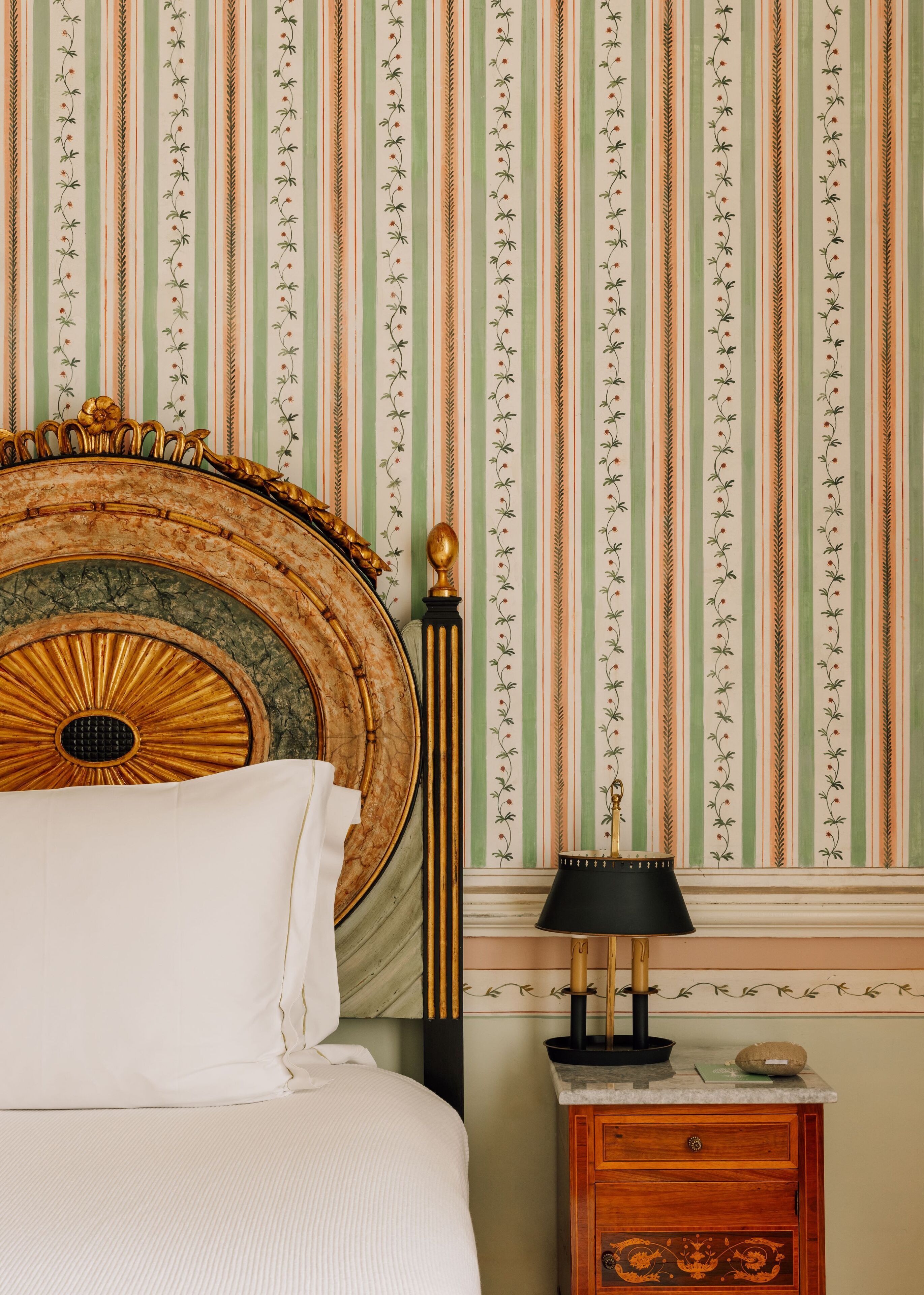 Quarto com cama, mesa de cabeceira, abajur e parede com papel de parede listrado.