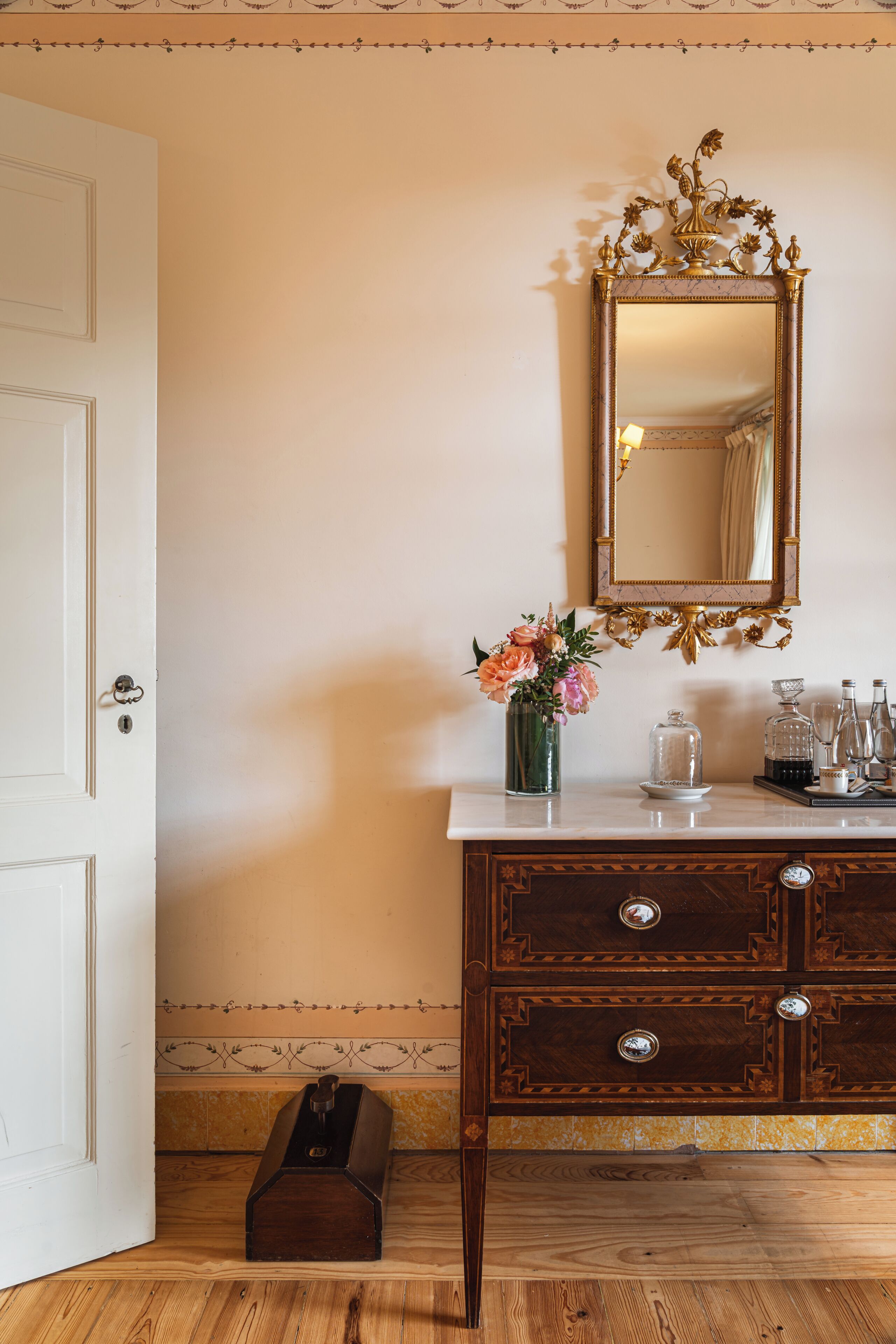 A room with a wooden dresser, a mirror with an ornate frame, a vase with flowers, and a small wooden box on the floor.