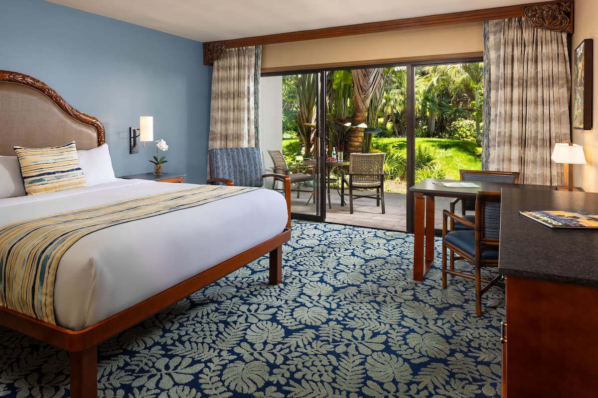 A hotel room with a large bed, a dining area with a table and chairs, and a view of a lush garden through a sliding glass door.