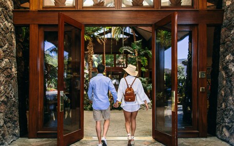 A couple walking through a wooden door into a building with a tropical garden view.