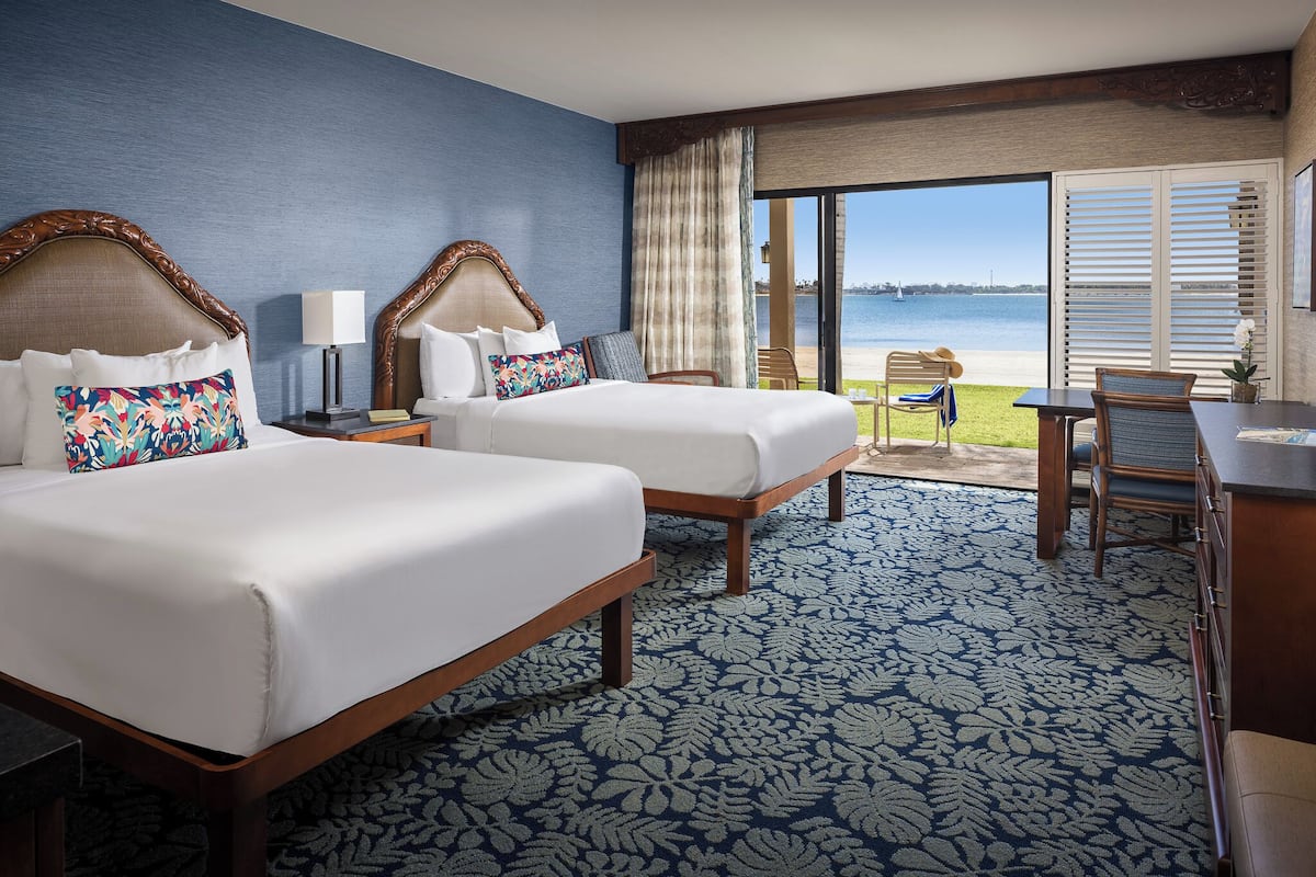 A hotel room with two beds, a large window with a view of the beach, and a balcony with a table and chairs.