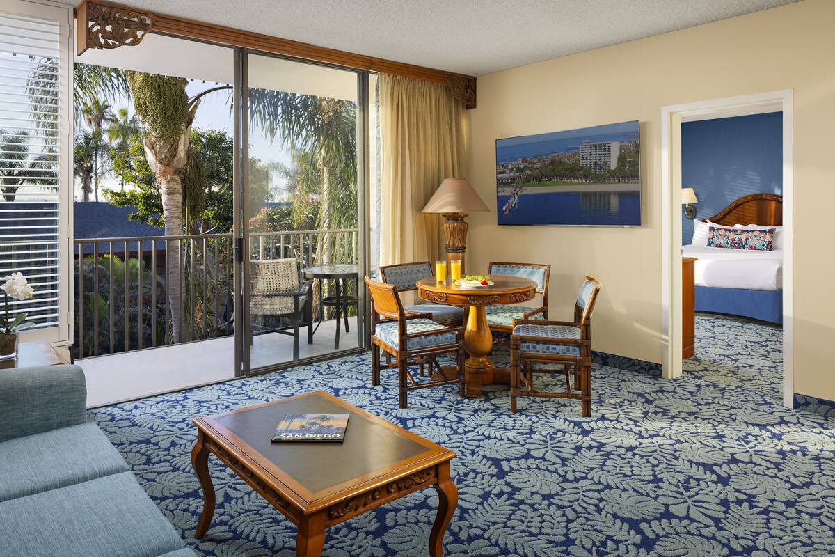 A hotel room with a balcony, a dining area with chairs and a table, a sofa, and a bed.