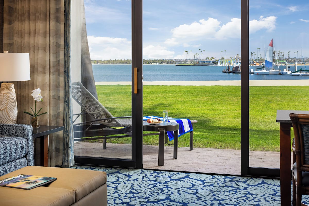 A room with a large glass door leading to a grassy deck with a table and chairs, overlooking a marina with boats and a clear sky.