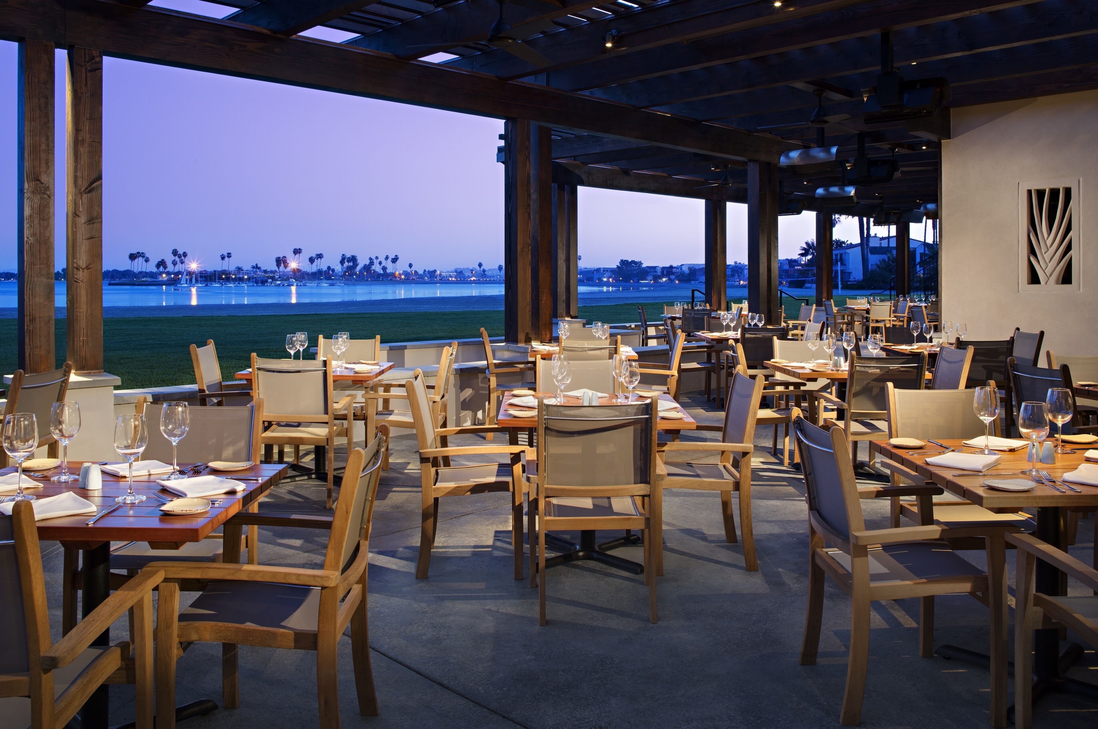 A restaurant with outdoor seating overlooking a body of water, featuring wooden tables and chairs, and a view of the coastline.