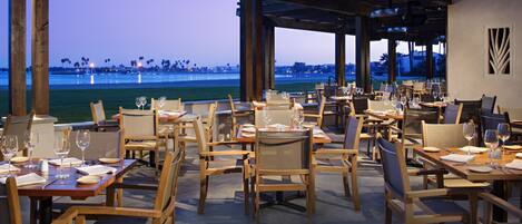 A restaurant with outdoor seating overlooking a body of water, featuring wooden tables and chairs, and a view of the coastline.
