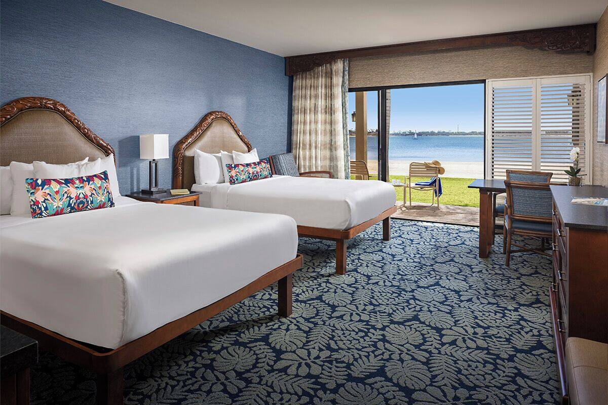 A hotel room with two beds, a large window with a view of the sea, a balcony with a table and chairs, and a patterned carpet.
