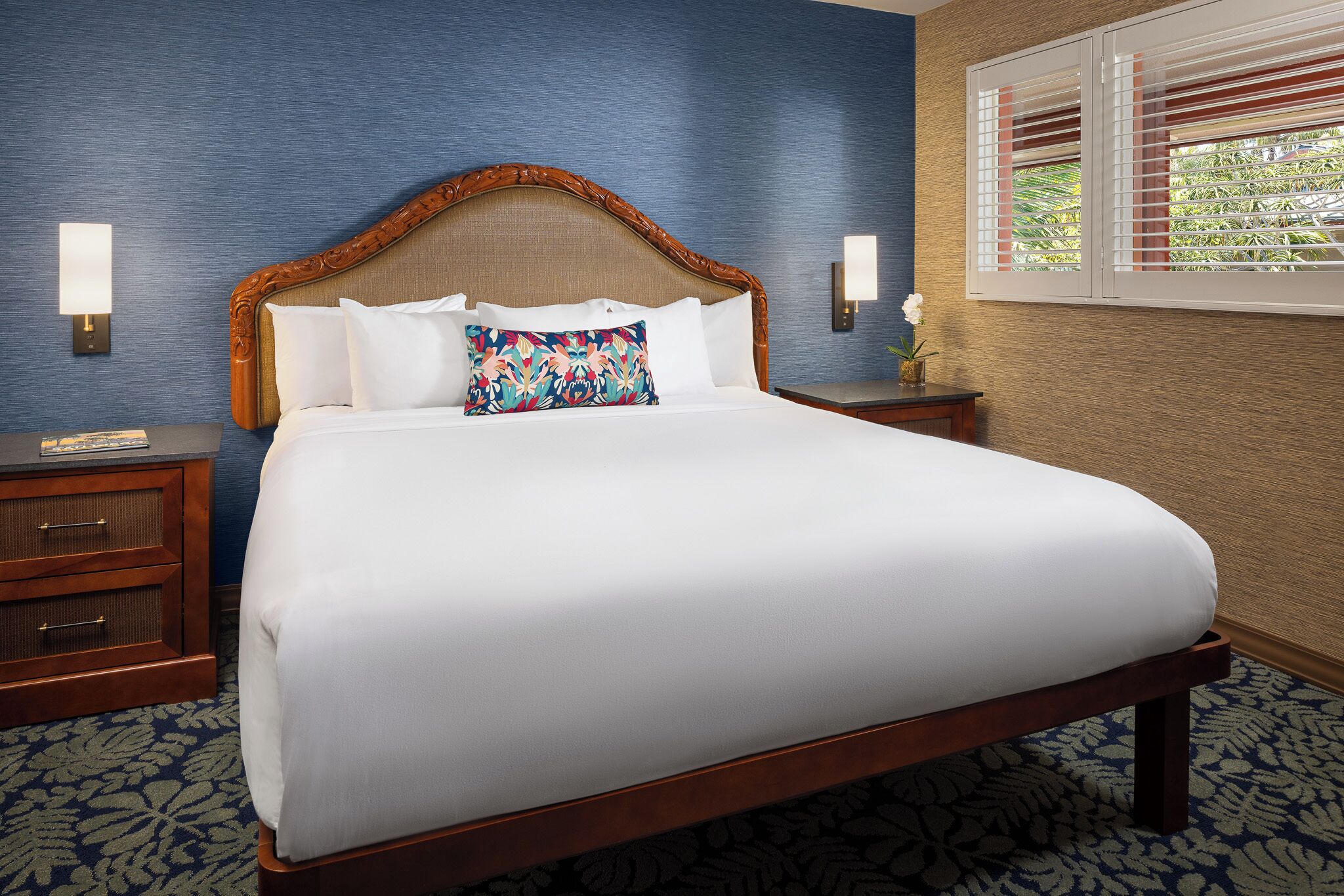A neatly made bed with a wooden headboard, white bedding, and a decorative pillow. There are two bedside tables with lamps and a nightstand with a telephone. The room has a patterned carpet and a window with blinds.
