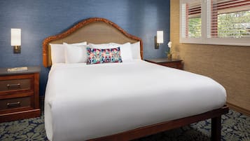 A neatly made bed with a wooden headboard, white bedding, and a decorative pillow. There are two bedside tables with lamps and a nightstand with a telephone. The room has a patterned carpet and a window with blinds.