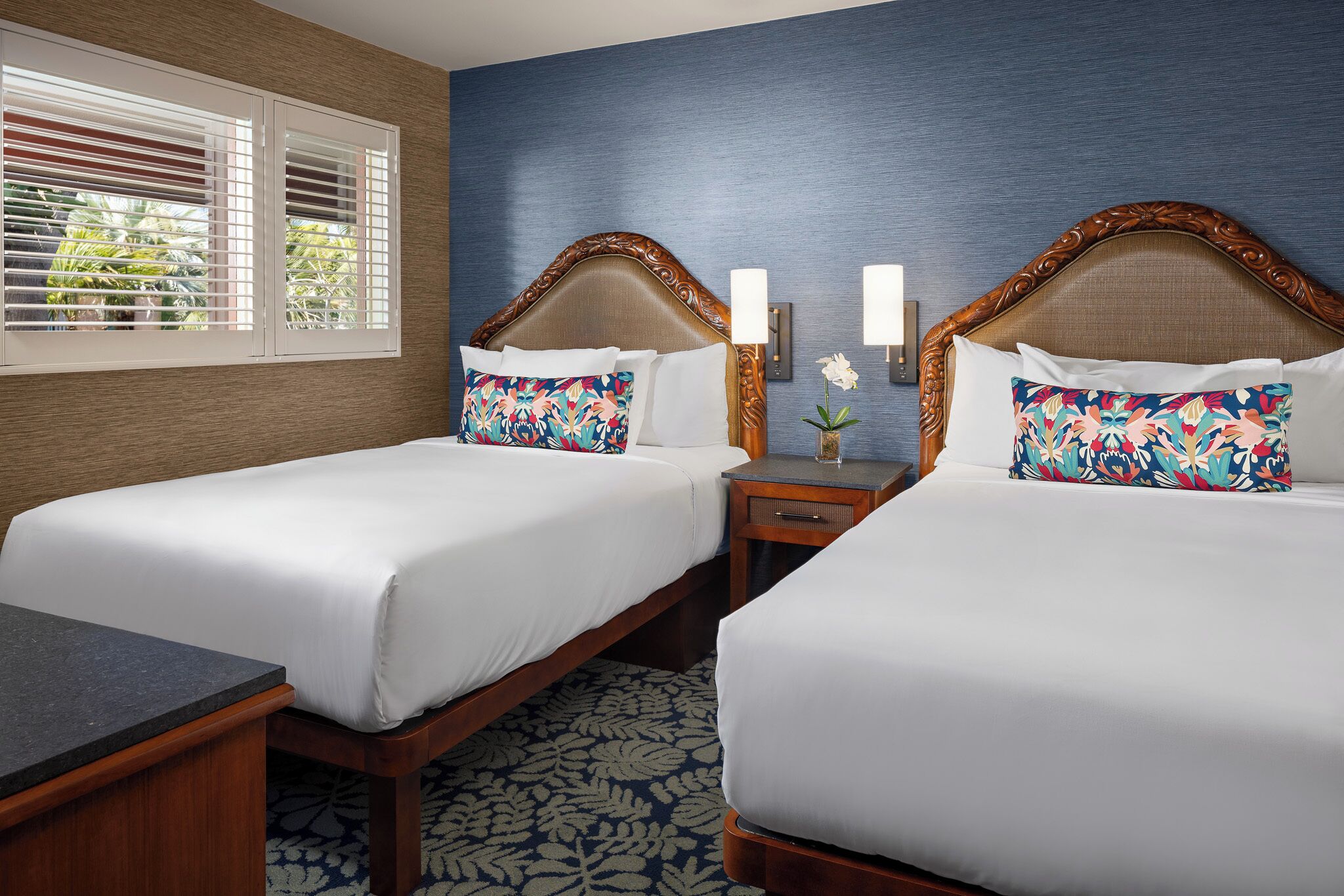 Quarto de hotel com duas camas, mesa de cabeceira, escrivaninha e janela com persianas.